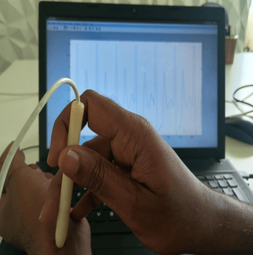 Hand holding a pulse sensor connected to a laptop displaying waveform data, representing AI-based Nadi Pariksha pulse diagnosis and digital health analysis