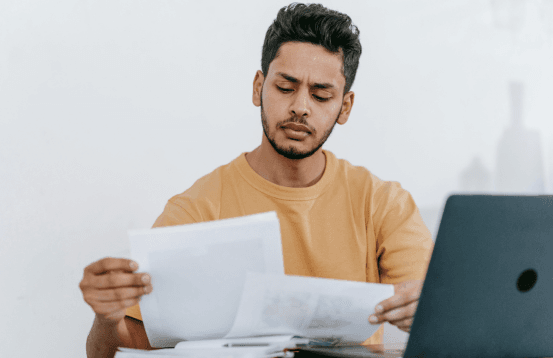 Young man reviewing documents with a focused expression while working on a laptop at a desk.